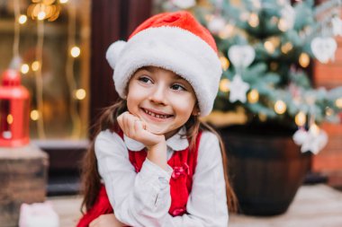 Noel Baba şapkalı, kırmızı elbiseli, süslü Noel ağacı olan bir evin verandasında oturan duygusal gülümseyen sevimli kız çocuğunun portresi. Noel Baba 'yı ve Noel tatillerini bekliyorum. Seçici odak.