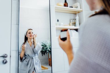 Young woman in bathrobe looking in the mirror and applying facial natural cosmetic clay mask on her face in bathroom. Cosmetic procedures for skin care at home. Beauty self-care. Selective focus.