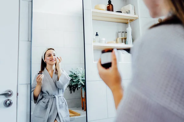 stock image Young woman in bathrobe looking in the mirror and applying facial natural cosmetic clay mask on her face in bathroom. Cosmetic procedures for skin care at home. Beauty self-care. Selective focus.