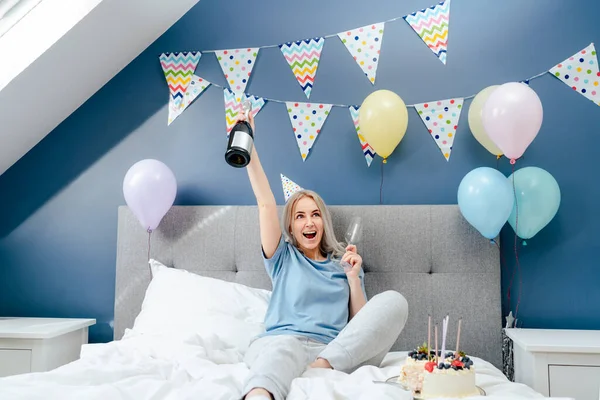 Happy emotional woman in pajama and party cap sitting on the bed with festive cake and champagne bottle in decorated bedroom. Time for yourself Celebration at home. Happy birthday concept