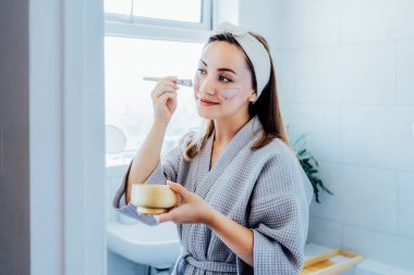 Young woman in bathrobe looking in the mirror and applying facial natural cosmetic clay mask on her face in bathroom. Cosmetic procedures for skin care at home. Beauty self-care. Selective focus.