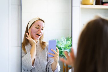 Yüzünde pembe kilden bir maske olan gülümseyen kadın ev banyosunda cep telefonuyla selfie çekiyor. Rahatlamanın ve güzellik tedavisinin tadını çıkarıyor. Güzellik blogcusu içerik yaratıyor. Seçici odak, boşluğu kopyala.
