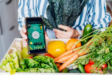 Çevrimiçi mobil Veganay Diyet Programı uygulaması ve sebzeli taze lahana kutusuyla telefonu elinde tutan bir yüz kadını yok. Sağlıklı beslenme, kilo verme. Yemek teslimatı, yemek tarifi.