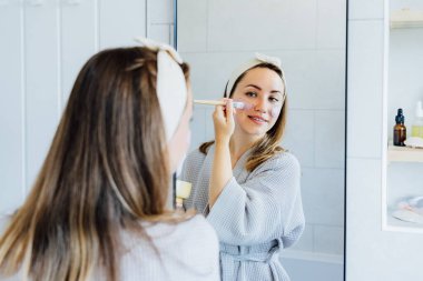 Young woman in bathrobe looking in the mirror and applying facial natural cosmetic clay mask on her face in bathroom. Cosmetic procedures for skin care at home. Beauty self-care. Selective focus