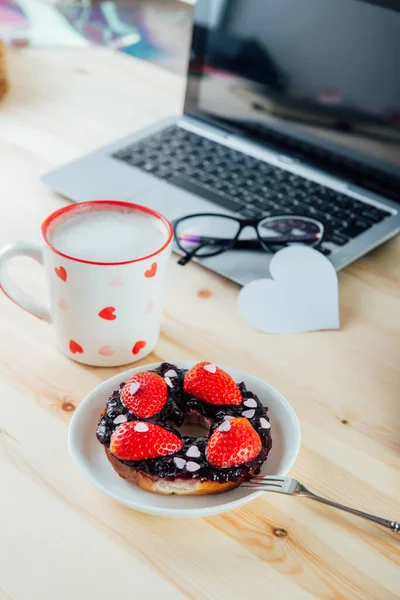 A tray with cup of latte coffee, bagel with strawberries and hearts, gift box and blank greeting card, rose flowers in vase on working place with laptop. Surprise for Valentine day. Selective focus.