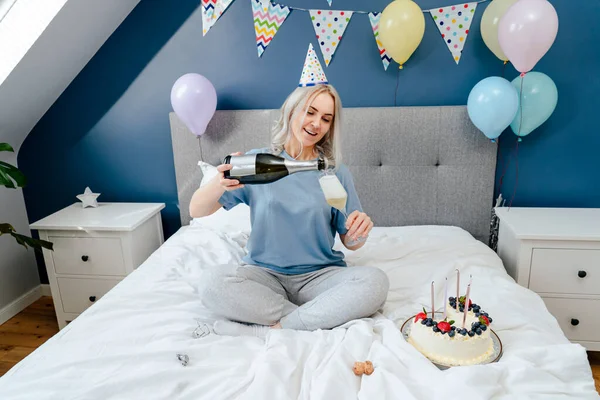 Happy emotional woman in pajama and party cap pouring champagne into glass, sitting on the bed with festive cake in a decorated bedroom. Time for yourself. Celebration at home. Happy birthday concept.