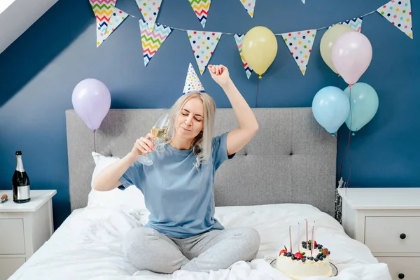 Happy emotional woman in pajama and party cap dancing on the bed with champagne glass in the hand and festive cake on the bed. Time for yourself Celebration at home. Happy birthday concept