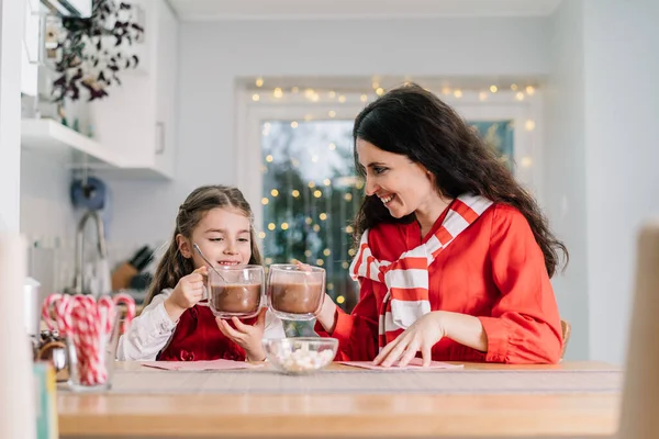 サンタの帽子の幸せな笑顔の娘の女の子と彼女のお母さんは 装飾されたモダンなキッチンに座って マシュマロでココアを飲んでいます 家族の休日だ クリスマスのお祭り気分 選択的焦点 — ストック写真
