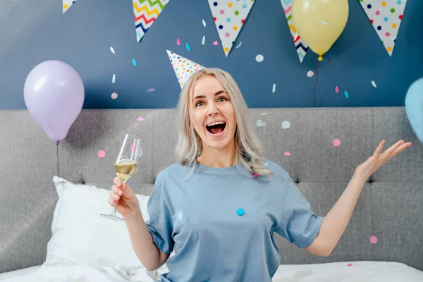 Happy emotional woman in pajama and party cap with champagne glass throwing confetti up while sitting on the bed in a decorated bedroom.Time for yourself Celebration at home. Happy birthday concept.