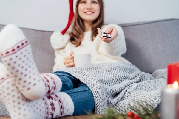 stock image Hand holding remote controller with blurred smiling woman in santa hat, jumper and warm socks sitting on comfortable couch watching movie, TV with christmas decoration atmosphere. Cozy winter concept.