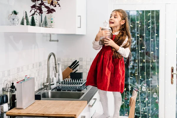 幸せな笑顔かわいい女の子の子供が椅子に立って 冬のクリスマス休暇のために装飾された棚の上にチョコレートのお祝いのクッキーと瓶に達しましたモダンなキッチン お菓子の食べ過ぎ — ストック写真