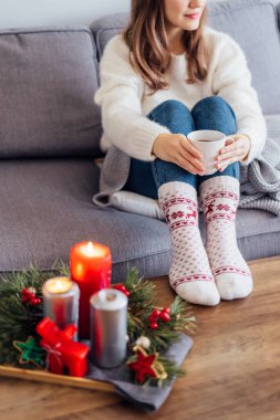 Süveter ve sıcak yün çoraplar giymiş bir kadın kanepenin üzerinde sıcak bir bardak sıcak içecek tutuyor ve sehpanın üzerindeki evde Hygge için Noel havasında mumlar yakıyor. Ev konforu. Kış ve Noel tatili. Dikey