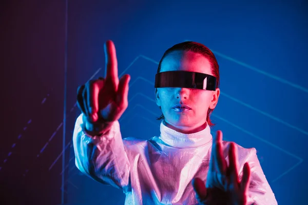 stock image Neon portrait of woman, dressed in futuristic holographic clothes and VR glasses touch space by finger. Cyberpunk girl with virtual touching pad. Neon blue and pink light. Virtual reality glasses.