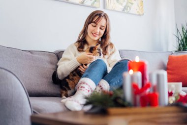 Hygge ev dekorasyonu için Noel kompozisyonu. Yanan mumlar, sehpadaki tepside çam ağacı dalları sıcak çay içen ve kanepede kedi besleyen dinlenen bir kadınla. Seçici odak