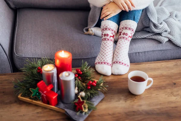 Female feet in xmas woolen socks, cup of hot drink and cozy Christmas mood candles composition for hygge home on the coffee table. Interior and home coziness concept. Winter and Christmas holidays.