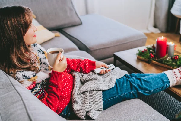 Relaxed woman in plaid and warm socks drinking hot tea with remote controller on sofa at home while watching movie, TV with christmas decoration atmosphere. Cozy and comfortable winter concept