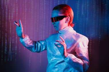 Geleceğe ait holografik giysiler ve gözlük takan kadın uzaya parmaklarıyla dokunuyor. Sanal dokunmatik bloknotu ve matris dijital numaraları olan siber punk kız neon mavisi ve pembe ışıkla arka plan