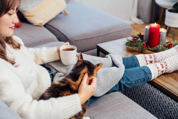 Relaxed woman with cup of hot drink petting cat on the sofa at home while watching movie, TV with christmas decoration atmosphere. Lady wear jumper and warm socks. Cozy and comfortable winter concept.
