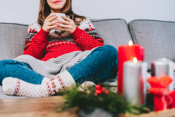 Cropped woman in ugly sweater and warm woolen socks on sofa holding cup of hot drink with cozy Christmas mood candles decor for hygge home on the coffee table. home coziness. Winter and Xmas holidays.
