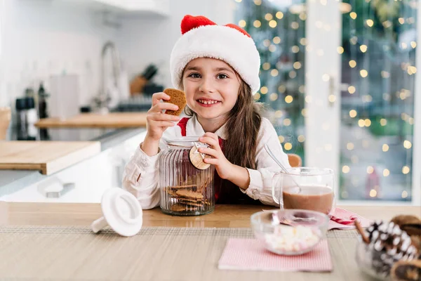 冬の休暇のために装飾されたモダンなキッチンに座っているサンタクロースの帽子のかわいい女の子の子供を笑顔とココアを飲みながら瓶からジンジャーブレッドクッキーに到達します 祭りの季節 — ストック写真