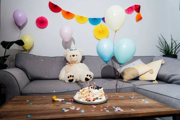 Messy table in living room after party. Broken birthday cake, confetti, balloons and decoration. Big teddy bear sitting on the sofa. Selective focus