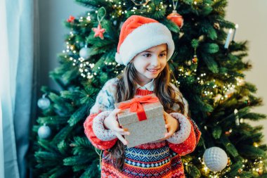 Noel Baba şapkalı, neşeli örgü süveterli mutlu bir kız Noel ağacının yanında duruyor. Noel konsepti. Noel Baba 'yı bekliyorum. Hediyeleri açıyorum. Seçici odak