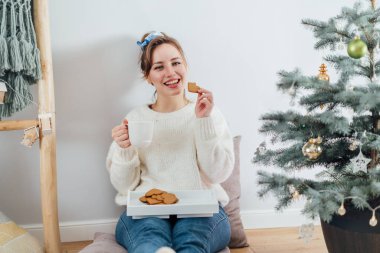 Rahat süveter giyen, sıcak çay içen, modern Scandi 'nin evindeki saksıdaki noel ağacının yanında dinlenen mutlu genç kadın. Çevre dostu sıcak kış tatili. Seçici odak.