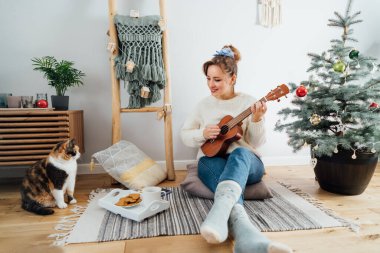 Ukulele gitar çalan genç bir kadın, modern İskandinavya evlerinde saksıdaki Noel ağacının yanında kedi evcil kedisiyle yerde dinlenirken. Şenlik konseri, müzik sanatı selamı. Seçici odak