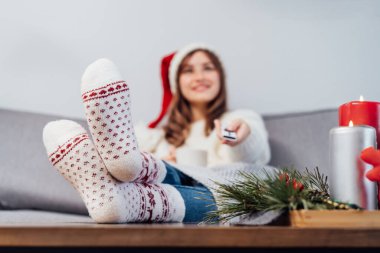 Yılbaşı süslemeli film izlerken, evde koltukta uzaktan kumandalı, Noel Baba şapkalı bulanık bir kadınla, Xmas yün çoraplarını giyip ayaklarına odaklan. Rahat kış konsepti