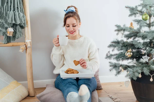 Kurabiyelerle sıcak çay içen, modern Scandi 'nin evindeki saksıdaki noel ağacının yanında dinlenen, rahat ve rahat genç kadın. Çevre dostu sıcak kış tatili. Seçici odak.