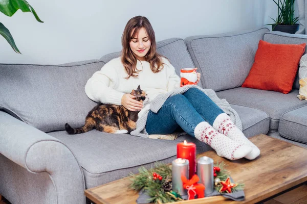 Relaxed woman with cup of hot drink petting cat on the sofa at home while watching movie, TV with christmas decoration atmosphere. Lady wear jumper and warm socks. Cozy and comfortable winter concept.