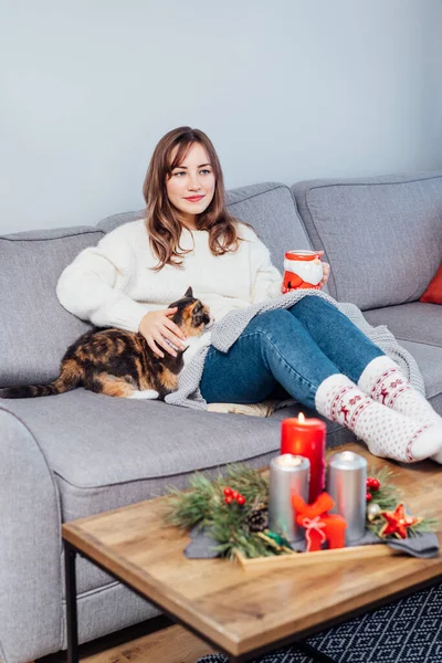 Relaxed woman with cup of hot drink petting cat on the sofa at home while watching movie, TV with christmas decoration atmosphere. Lady wear jumper and warm socks. Cozy and comfortable winter concept