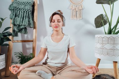 Sağlıklı genç bir kadın evde meditasyon yapıyor, kulaklık takıyor, modern Scandi 'nin evindeki minderde lotus pozisyonunda oturuyor. Meditasyon, rahatlama, gözler kapalı. Seçici odak.