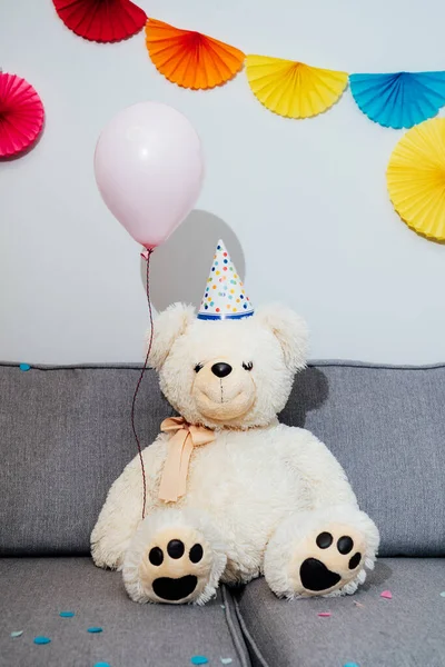 Big Teddy bear with balloon in party cap and sitting on the gray sofa in decorated room. Birthday or festive holiday vertical background. Selective focus, copy space