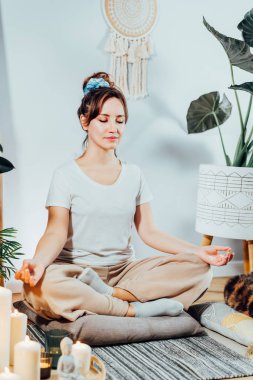 Genç sağlıklı bir kadın evde meditasyon yapıyor, modern Scandi 'nin evindeki yerde lotus pozisyonunda oturuyor. Meditasyon, rahatlama, gözler kapalı. Dikey kart. Seçici odak.