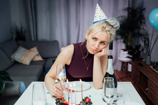 Bored, sad attractive woman drinking champagne while celebrating birthday at home, sitting alone at served table with cake, keeping hand under chin, looking away, dreaming. Selfparty. Selective focus.
