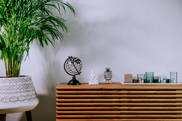 Scandinavian home interior with decorative accessories standing on a wooden cabinet. Minimalist design in interior of room with green plants and white wall with copy space. Biophilia style.
