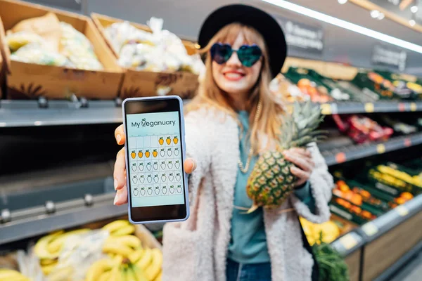 Close up phone with online mobile app of Veganuary diet calendar. Stylish fashion woman holding pineapple in the supermarket store during selecting products. Healthy eating, go vegan. Selective focus