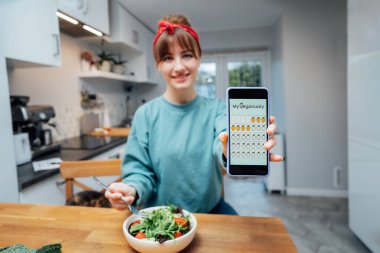 Focused phone with active online mobile app of Veganuary diet calendar in womans hand, who is eating fresh green salad on kitchen. Healthy vegetarian and vegan food diet planning. Selective focus