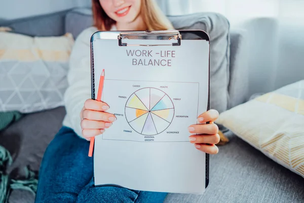 Close up drawn work-life balance wheel diagram holding by smiling woman sitting on the sofa at home. Self-reflection and life planning. Coaching tools. Finding Balance in Your Life. Selective focus.