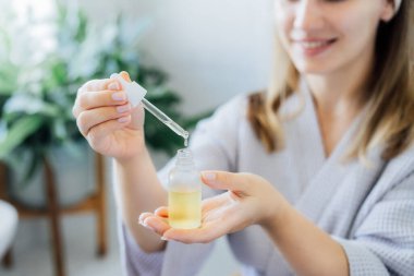 Close up young woman in bathrobe holding mockup bottle and dropper with hyaluronic acid serum in her hands. Skin hydrating. Cosmetic spa procedures. Beauty self-care at home. Selective focus