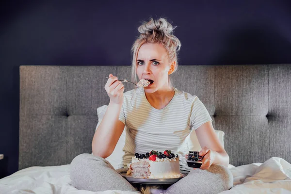 Emotionally stressed, shocked woman eating cake and holding a remote control while watching TV in bed. Scary horror movie or terrible news, scare in her eyes Overeating, Jamming of negative emotions.