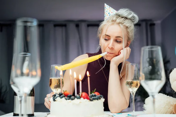 Sad attractive woman in party cap blowing in birthday pipe while celebrating birthday at home, sitting alone at served table with cake, keeping hand under chin, looking away, dreaming. Bored party