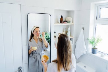 Young woman in bathrobe looking in the mirror and applying natural cosmetic clay mask on her face in bathroom. Cosmetic procedures for teens skin care at home. Beauty self-care. Selective focus