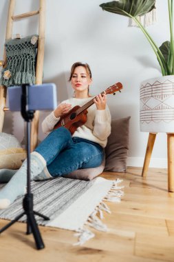 Ukulele gitar çalan genç bir kadın, modern Scandi iç mekanında telefonla müzik videosu kaydediyor. Blog yazarı şarkı söylüyor, içerik yaratıyor. Çevrimiçi öğrenme, öğretme, performans, eğlenceli konsept.