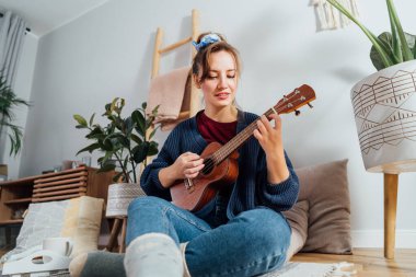 Young woman playing ukulele guitar while relaxing on floor cushion in modern scandinavian home interior.Musical Hobby, home fun concept. Resolution to learn new skills. Selective focus.