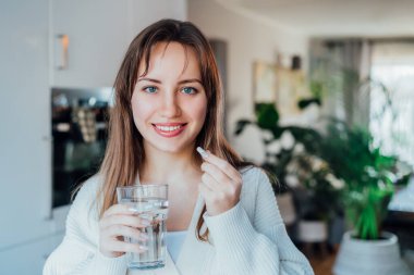 Young smiling woman holding pill and glass of water at home kitchen. Positive lady takes daily medicine antioxidant diet vitamin supplements for beauty skin hair health care medicament concept