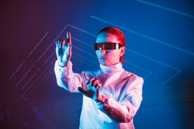 Neon portrait of woman, dressed in futuristic holographic clothes and glasses touch space by finger. Cyberpunk girl with virtual touching pad. Neon blue and pink light. Virtual reality glasses