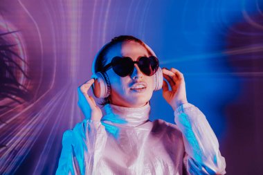Portrait of smiling woman in hear shaped sunglasses and headphones in neon light. Music lover. Silent disco. Dj in holographic clothes on the background of music vibes background. Futuristic Party
