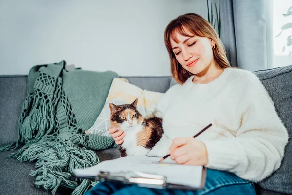 Relaxed young woman drawing work-life balance wheel sitting on the sofa with cat pet at home. Self-reflection and life planning. Coaching tools. Finding Balance in Your Life. Selective focus.